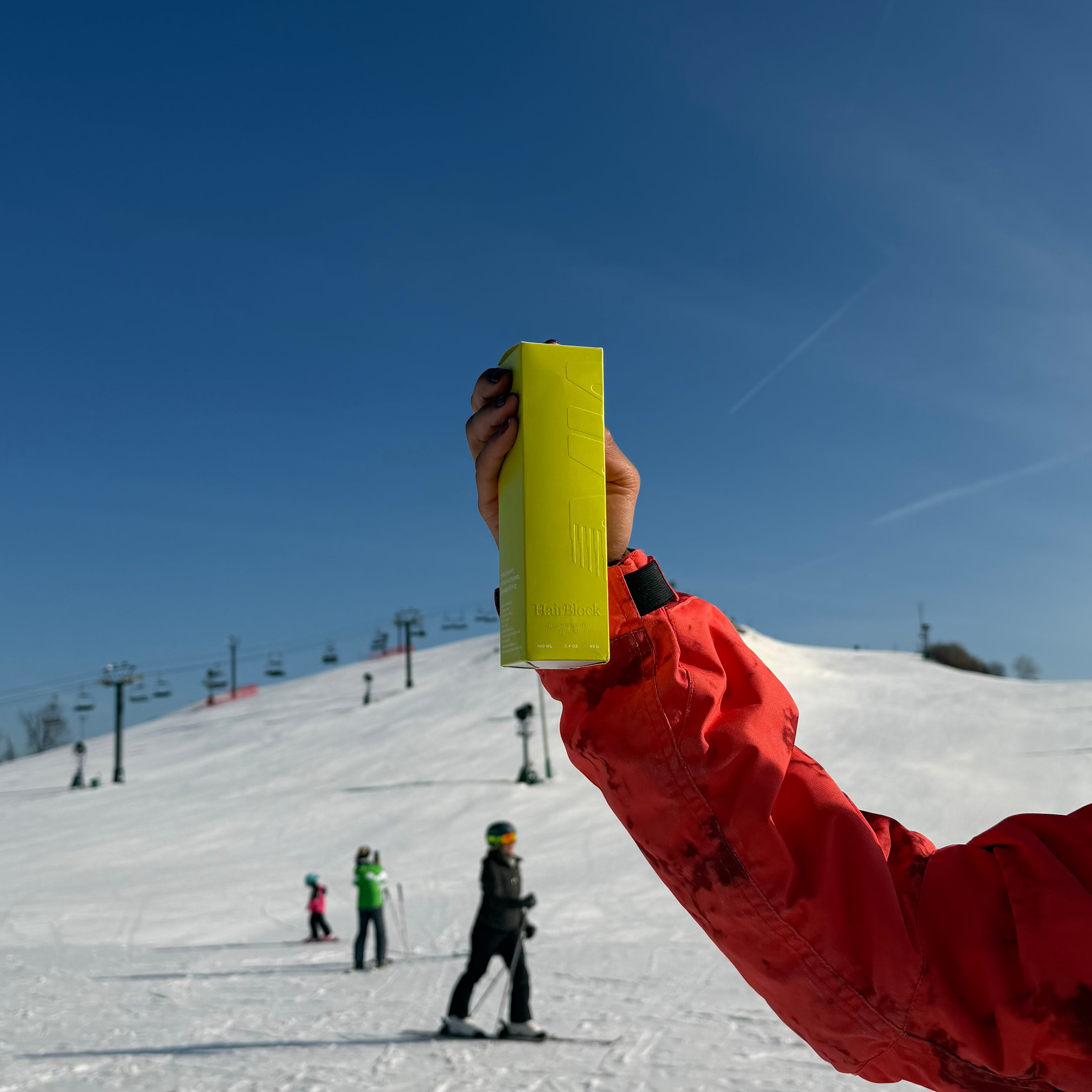 Person in red jacket holding a yellow object on a snowy slope with skiers in the background